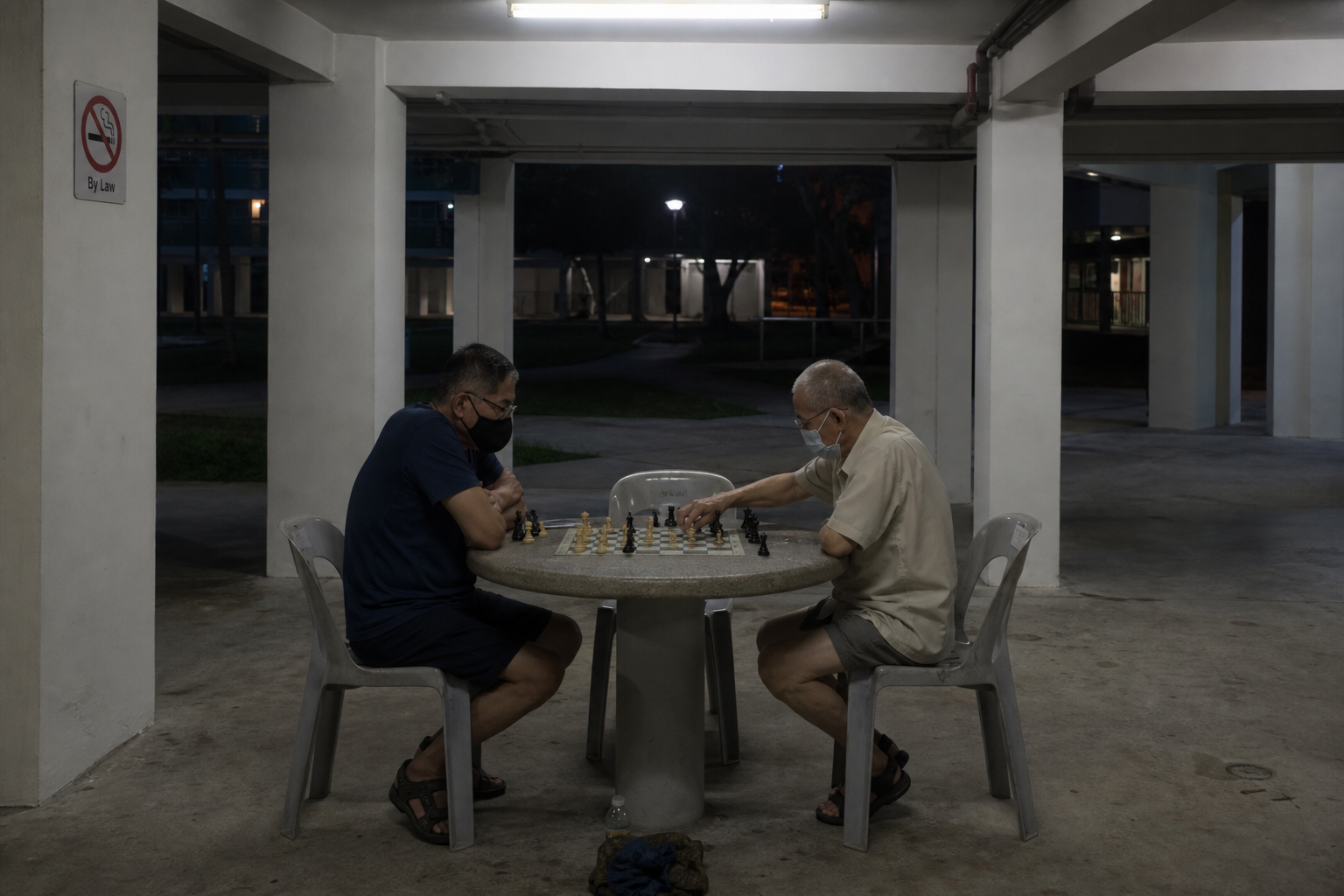 Void-deck chess table — pieces mid-game, plastic chairs, fluorescent tube light above